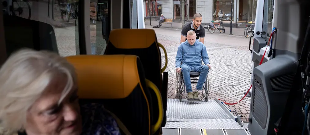 Inifrån en sjuktransport, en äldre kvinna sitter i bilen , en yngre man i rullstol rullas in bak i bilen.
