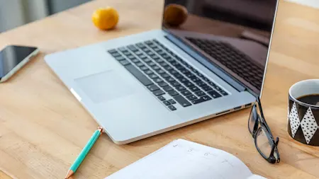 En laptop, en kaffekopp, en agenda och en clementin allt på ett bord.