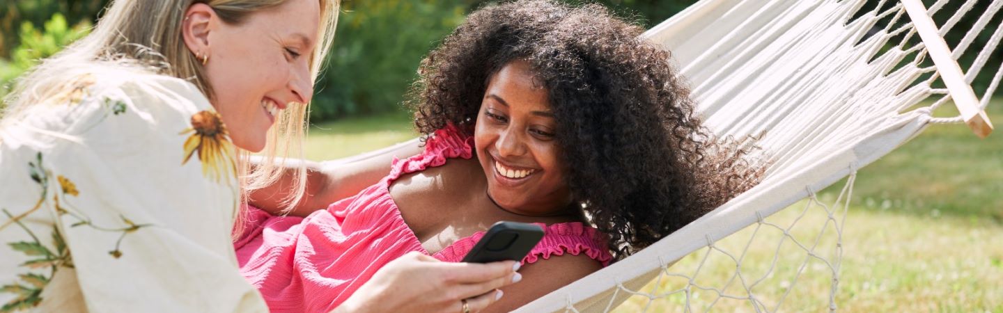 Två kvinnor i somriga kläder utomhus tittar på en mobiltelefon. En av dem sitter på gräset och den andra ligger i en hängmatta.