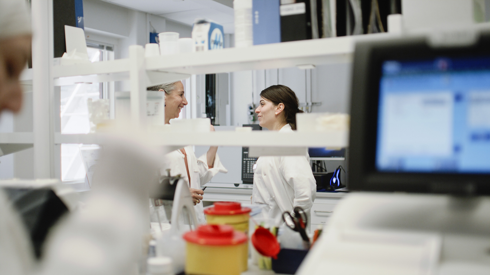 Två kvinnor i vita rockar ler mot varandra i ett laboratorium. Framför dem står hyllor och en dator.