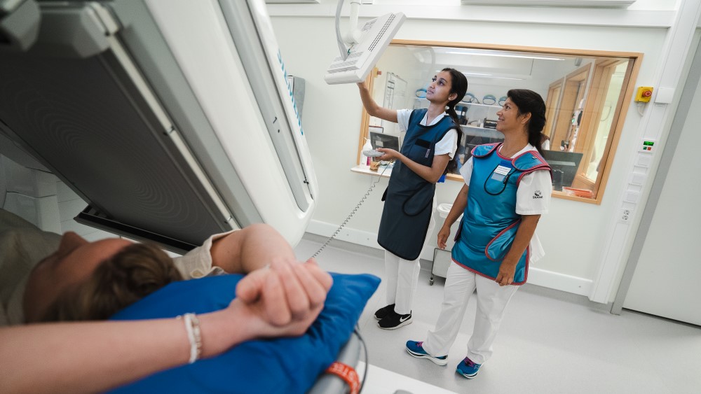 Två kvinnor i sjukhuskläder och skyddsväst för röntgen tittar på en skärm. En person ligger under en maskin och blir röntgad.