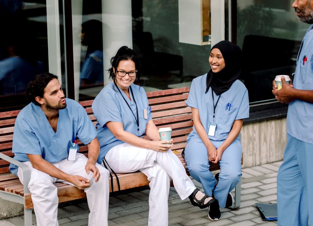 Fyra personer i sjukhuskläder pratar med varandra utomhus. Tre av dem sitter på en bänk medan en står. Några av dem håller i kaffemuggar och de se glada ut.