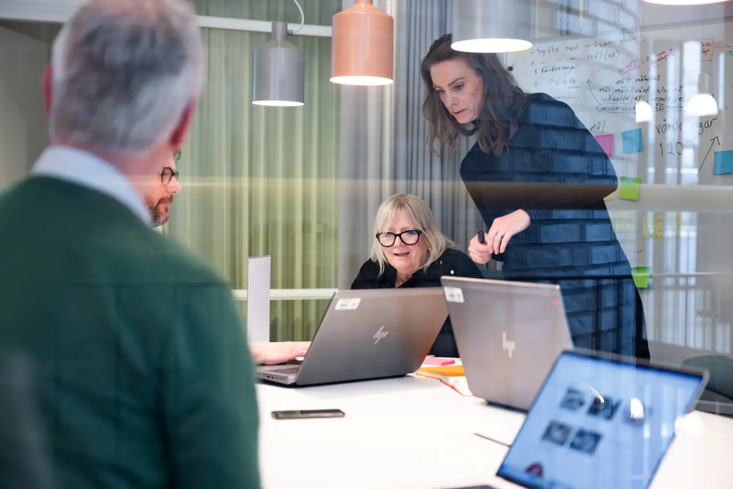 Fyra personer sitter och står kring ett bord i kontorsmiljö. På bordet står flera öppna laptops.