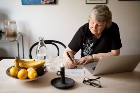 En äldre kvinna sitter vid ett bord och antecknar på ett papper. Framför henne står en öppen laptop. På bodet ligger ett par glasögon och det står en fruktskål med citroner och bananer på bordet.
