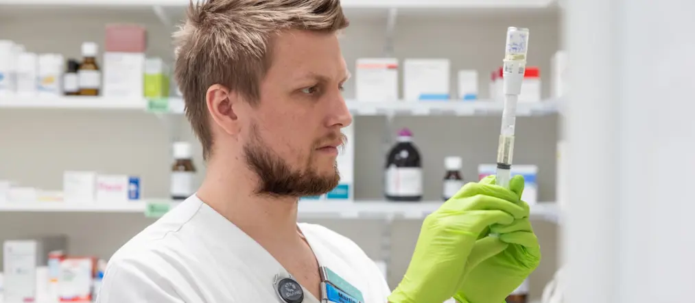 En manlig sjuksköterska tittar koncentrerat på ett läkemedel som han håller i med plasthandskar. I bakgrunden syns hyllor med fler läkemedel i flaskor och lådor.