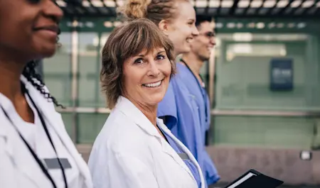 En person som tittar i kameran leendes iklädd en vit vårdrock med stetoskop runt halsen.