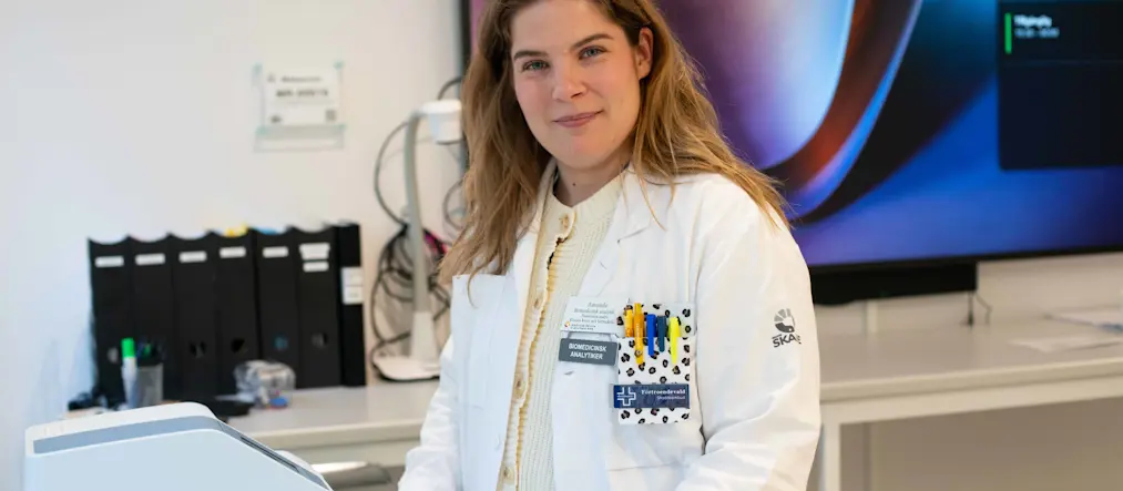 Amanda Gråberg, biomedicinsk analytiker, klädd i vita vårdkläder, står i ett labb, tittar in i kameran.
