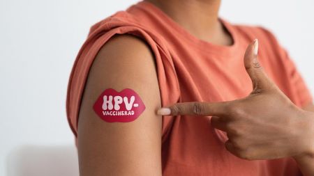 Kvinna pekar på sin överarm där det sitter ett plåster i form av en pussmun med texten HPV vaccinerad