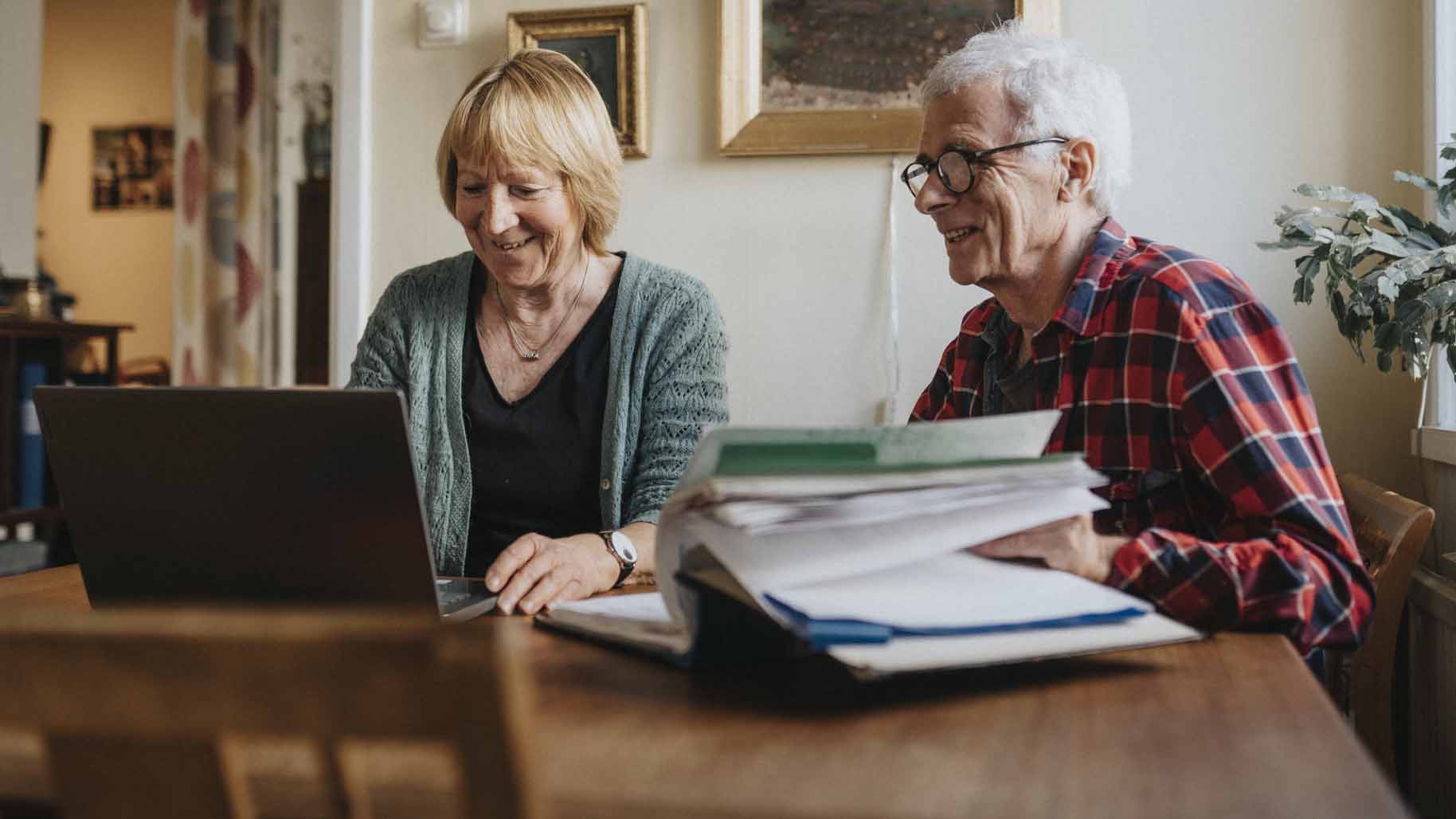 ett äldre par, kvinna och man, sitter vid ett bord och tittar på en laptop, ler.