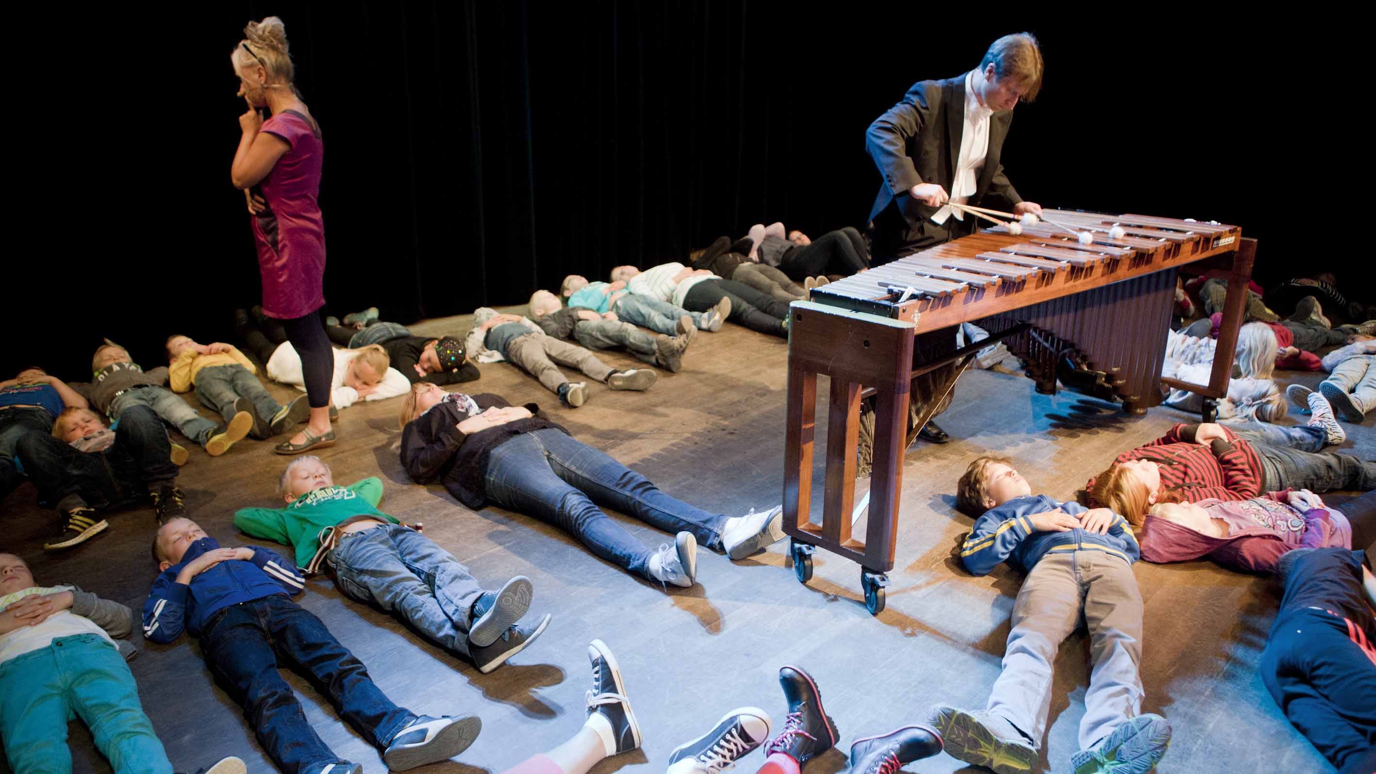 barn ligger utsträckta på ett trägolv, i mitten står en marimbaspelare och spelar på sin marimba. I utkanten av bilden en kvinna klädd i rosa.